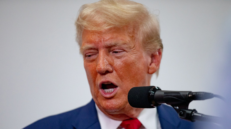 Donald Trump's face looks greasy and red as he speaks into a microphone and glances to the side.