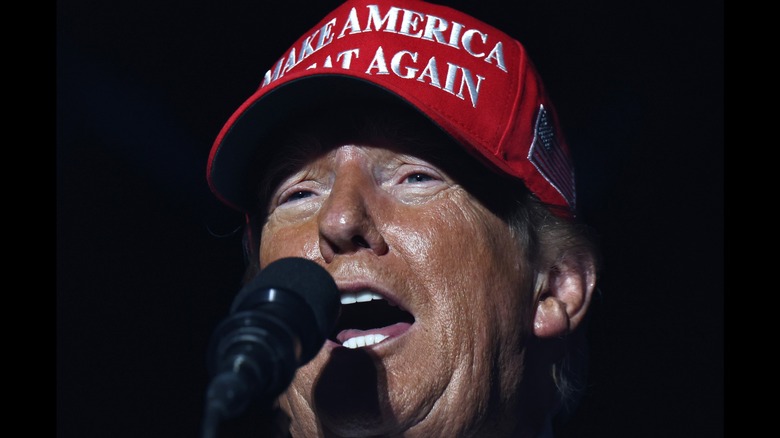 A sweat Donald Trump wears a MAGA hat and speaks into a microphone during a rally in Coachella.