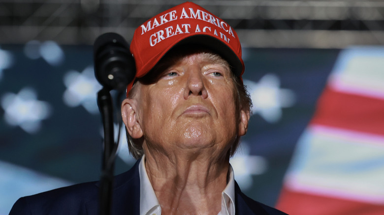 A frowning Donald Trump wears a MAGA hat and sweats during a rally.