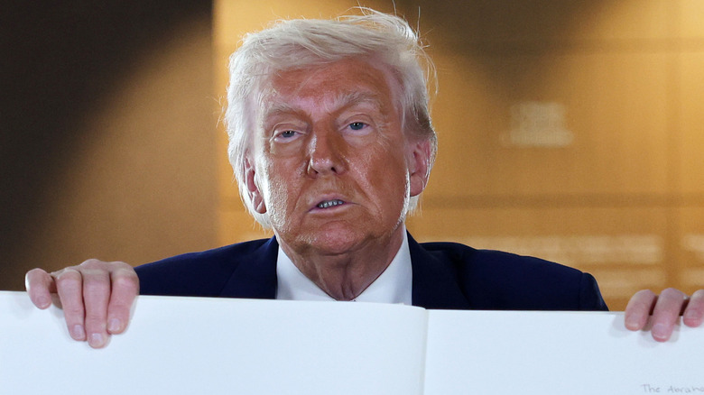 A tired-looking Donald Trump holds up a large guest book.
