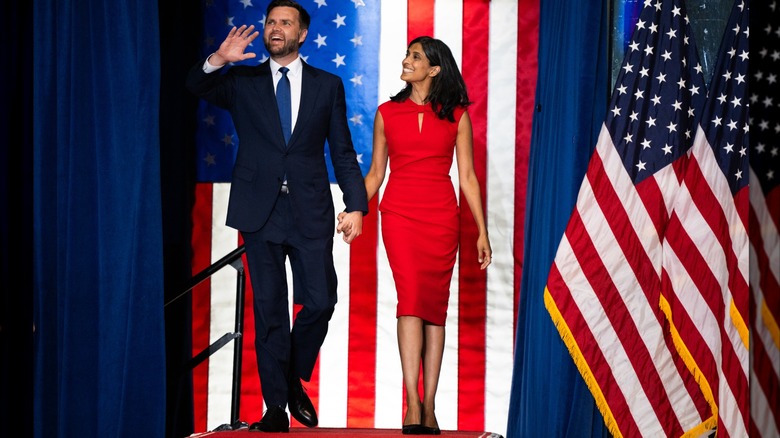 Usha Vance and JD Vance walking in front of American flags, she in a tight red dress