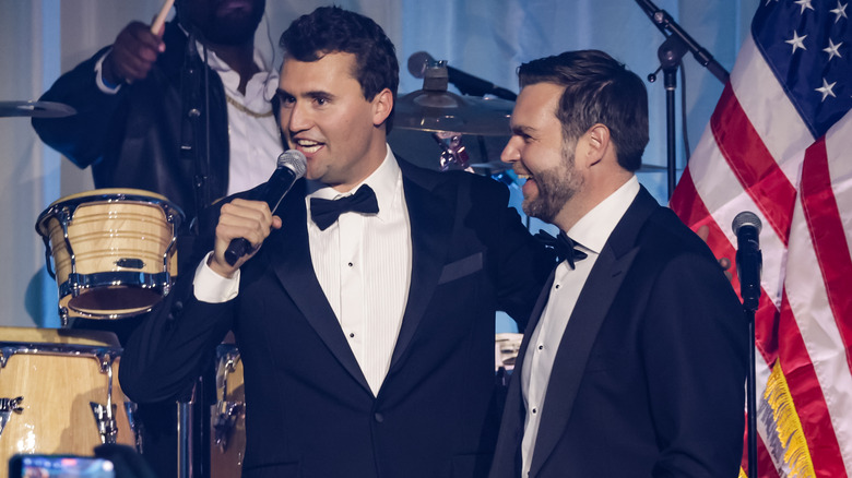 Charlie Kirk smiling beside JD Vance onstage with a mic while both wearing tuxedos