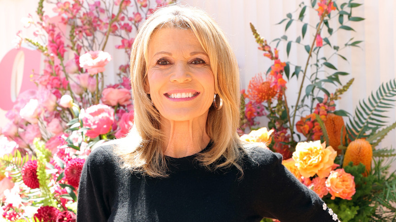 Vanna White blonde hair down wearing a full face of makeup and a black sweater with flowers behind her