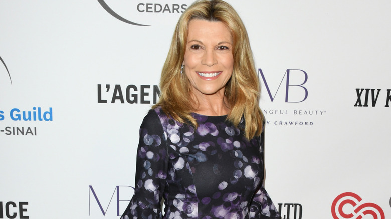 Vanna white smiling with blonde hair down full face of makeup purple dress.