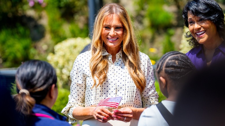 Melania Trump holding a small American flag and smiling surrounded by people (2025)