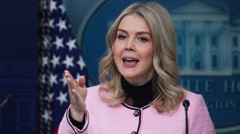 Karoline Leavitt giving a press briefing in a pink blazer while gesturing with her hand