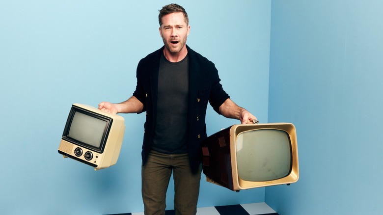 Luke Macfarlane posing with a couple of old TVs in his hands