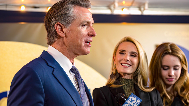 Gavin Newsom speaking to reporters while Jennifer Siebel looks on