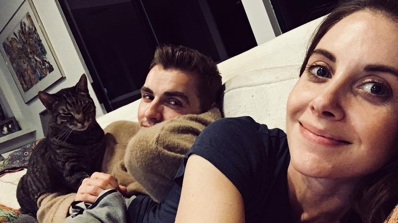 Alison Brie and Dave Franco stretched out on a couch with one of their cats