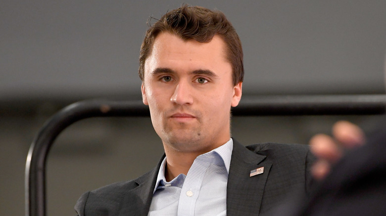 Charlie Kirk sitting at an event with a neutral expression while wearing a dark grey suit