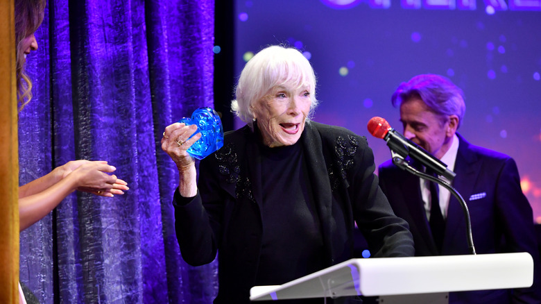 Shirley MacLaine smiling on stage and holding a blue heart-shaped award in 2025
