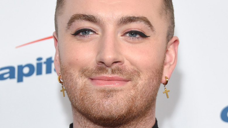 Sam Smith smiling wearing cross earrings and winged eyeliner