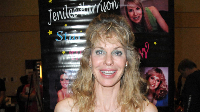 Jennilee Harrison posing in a sleeveless dress, smiling in front of a poster featuring old pictures of herself