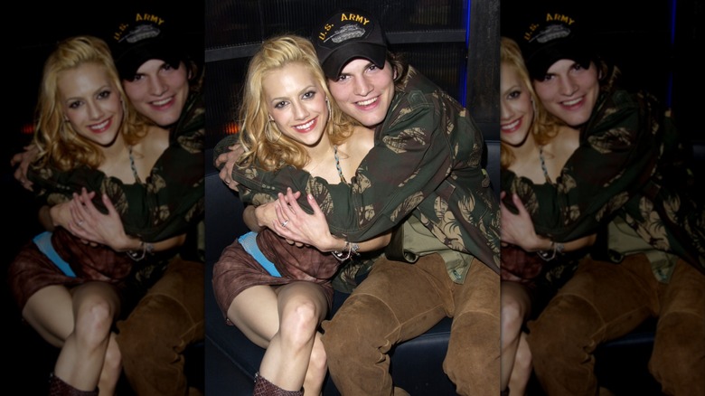 Brittany Murphy and Ashton Kutcher smiling as they sit with their arms wrapped around each other