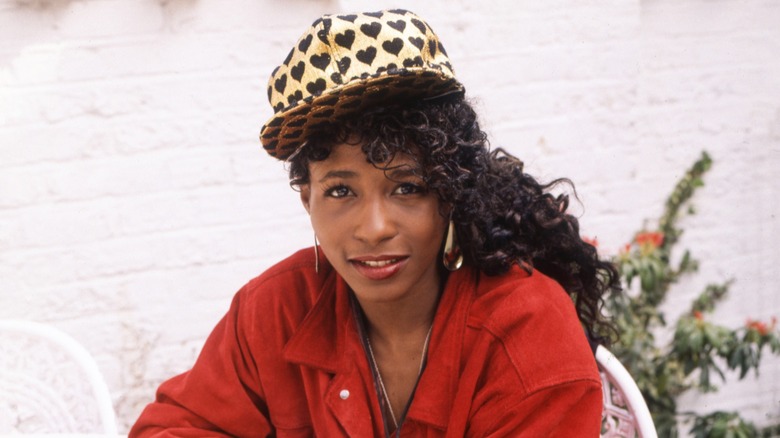 Sinitta sitting down wearing a red jacket and black-and-gold heart-print baseball cap.
