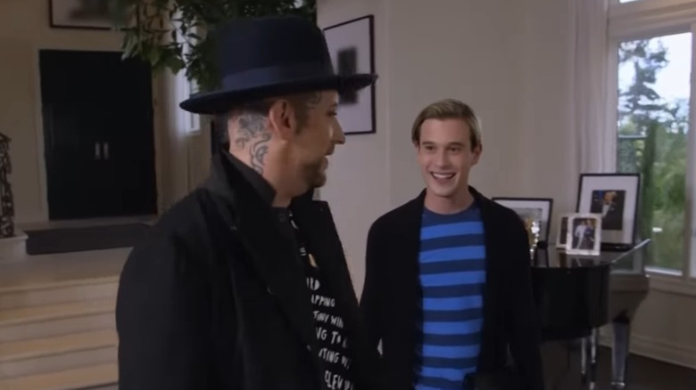 Boy George looking at Tyler Henry as they walk past a piano with framed photos on top