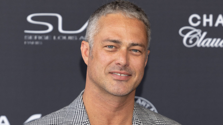 Taylor Kinney smiling while posing at an event in a grey check blazer