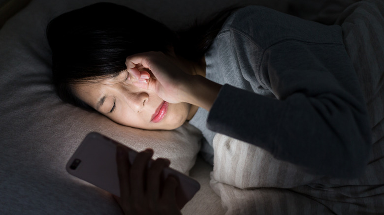 Woman laying in bed rubbing eyes while using mobile phone