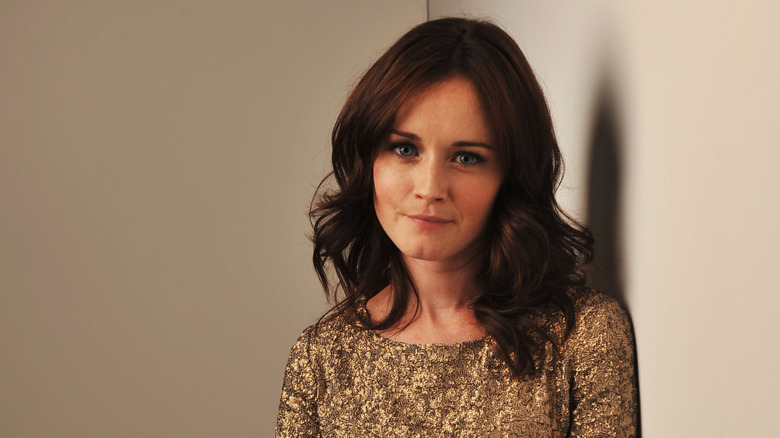 Alexis Bledel looking into the camera wearing a gold dress in front of a beige wall.