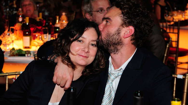 Johnny Galecki hugs Sara Gilbert around the neck and kisses her on the head while they sit together at a table.