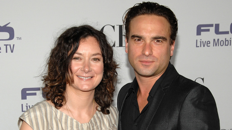 Sara Gilbert wears a beige striped shirt and smiles next to Johnny Galecki, who wears a black jacket and matching shirt.