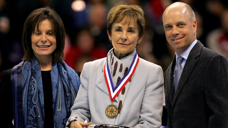 Olympic figure skater Scott Hamilton stands next to two women smiling as he is inducted into the figure skating Hall of Fame.