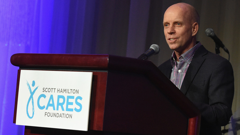 Former figure skater Scott Hamilton stands at a podium speaking at an event for his cancer foundation