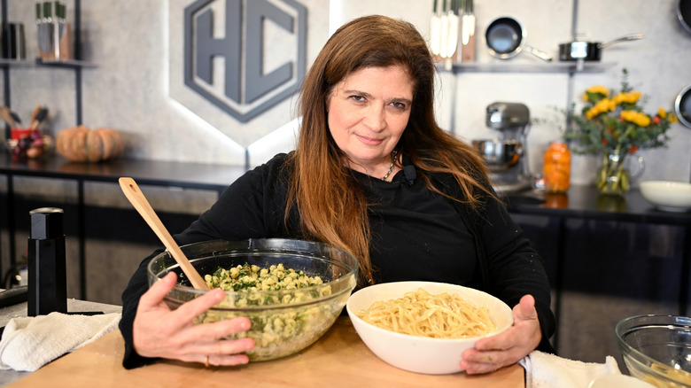 Alex Guarnaschelli poses mouth closed holding two bowls of food wearing a black chef coat