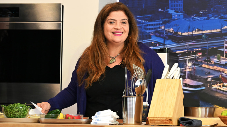 Alex Guarnaschelli smiles on set with vegetables and cooking utensils in front of her.