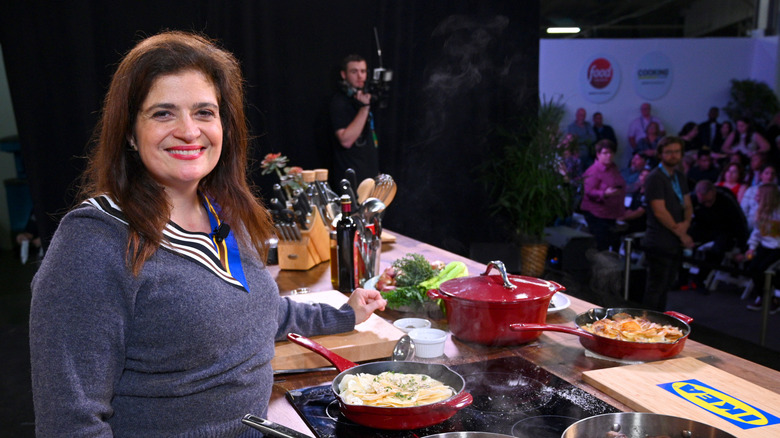 Alex Guarnaschelli smiles on set in front of a stove cooking in a blue sweater