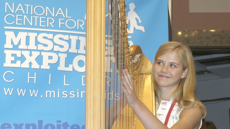A teenage Elizabeth Smart playing the harp in front of a poster for the National Center for Missing and Exploited Children