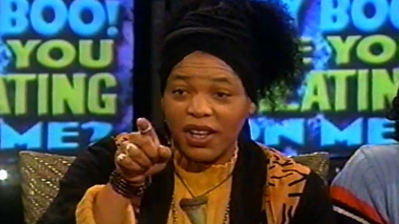 Miss cleo gesturing directly toward the camera. The background displays bold text in bright colors.