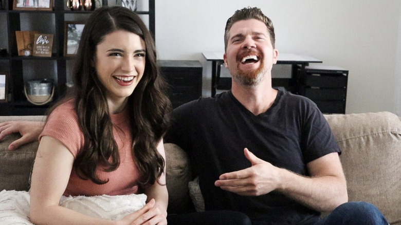 Beau Wirick and his wife Daniela Bobadilla sit on a couch in a living room next to each other laughing.