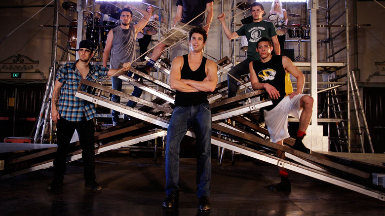 Adam Garcia posing with the cast of Tap Dogs onstage with his arms crossed in a black tank top and jeans