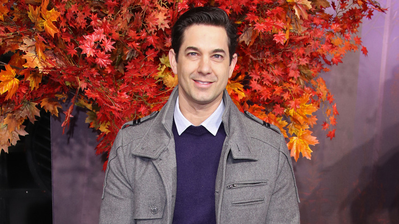 Adam Garcia smiling in front of autumn leaves wearing a gray jacket