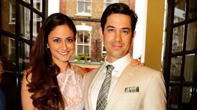 Adam Garcia smiles in a tan suit next to his wife in a pink dress outside in London