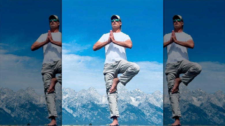 Josh Holloway does a yoga pose barefoot outside with a snowy mountainscape behind him