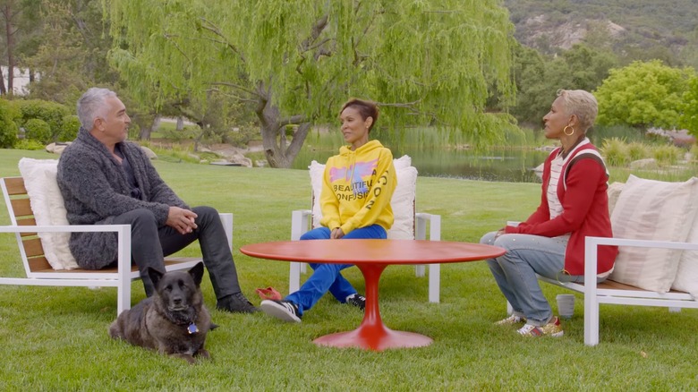 Cesar Millan, a dog, Jada Pinkett Smith, and Adrienne Banfield-Norris sitting around a red table outside in front of a willow tree