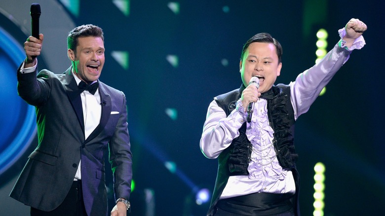 Ryan Seacrest raising a microphone as William Hung sings into his own mic and raises a fist