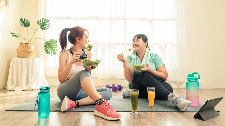 two women eating salad on a yoga mat