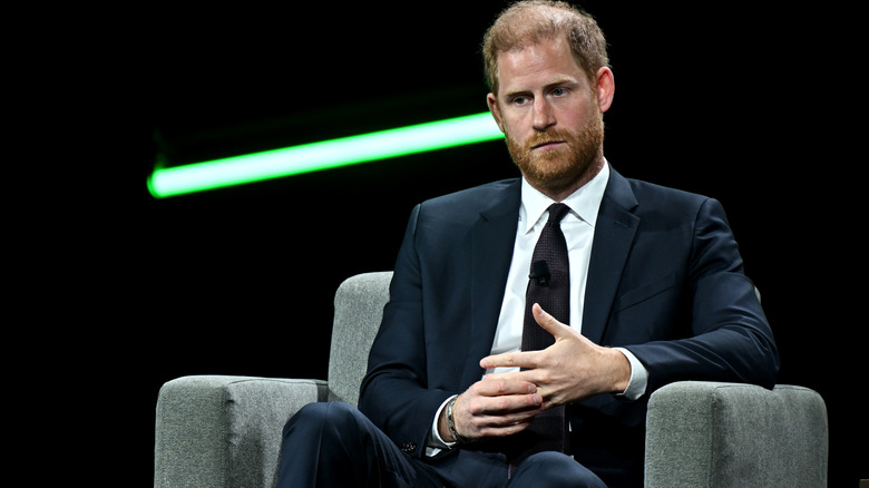 Prince Harry looking weary while sitting in a grey chair in a black suit
