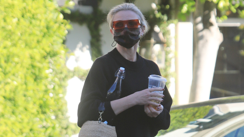 Cameron Diaz photographed on street wearing sunglasses, a face mask, and carrying a water bottle, an iced drink, and handbag