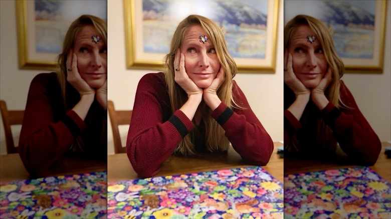 Jane Sibbett with a puzzle piece on her forehead, leaning on her elbows with a puzzle on the table before her.