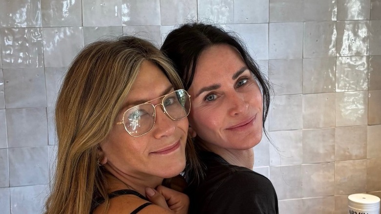Jennifer Aniston cozied up to Courteney Cox in a white walled and white tiled bathroom.