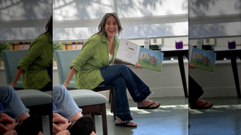 Maggie Wheeler leaning forward in a seat, holding out a children's book.