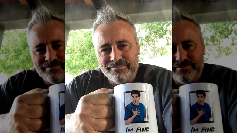 Matt LeBlanc outside, leaning over his phone, holding a Friends themed mug.