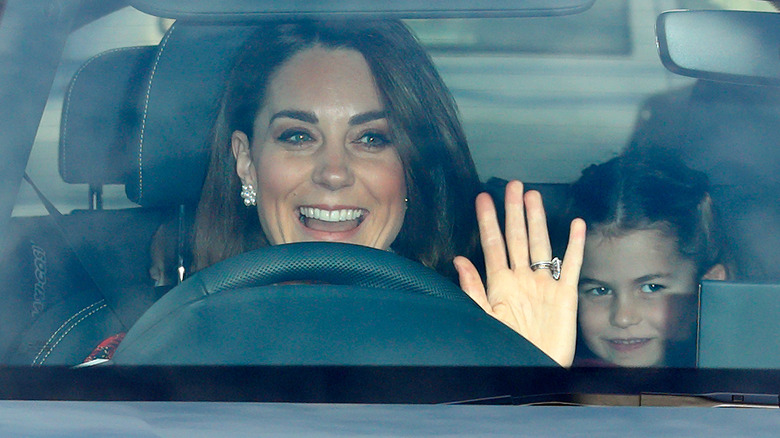 Princess Catherine waving at the camera from behind the wheel with a smiling Princess Charlotte in the back seat