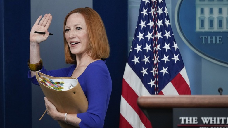 Jen Psaki waving as she leaves the briefing room, binder under her arm