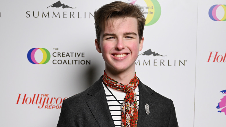Iain Armitage smiling while posing at an event in a black blazer, stripy top, and red patterned scarf
