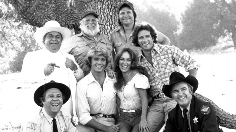 The cast of The Dukes of Hazzard pose outside next to each other smiling in a black and white photo